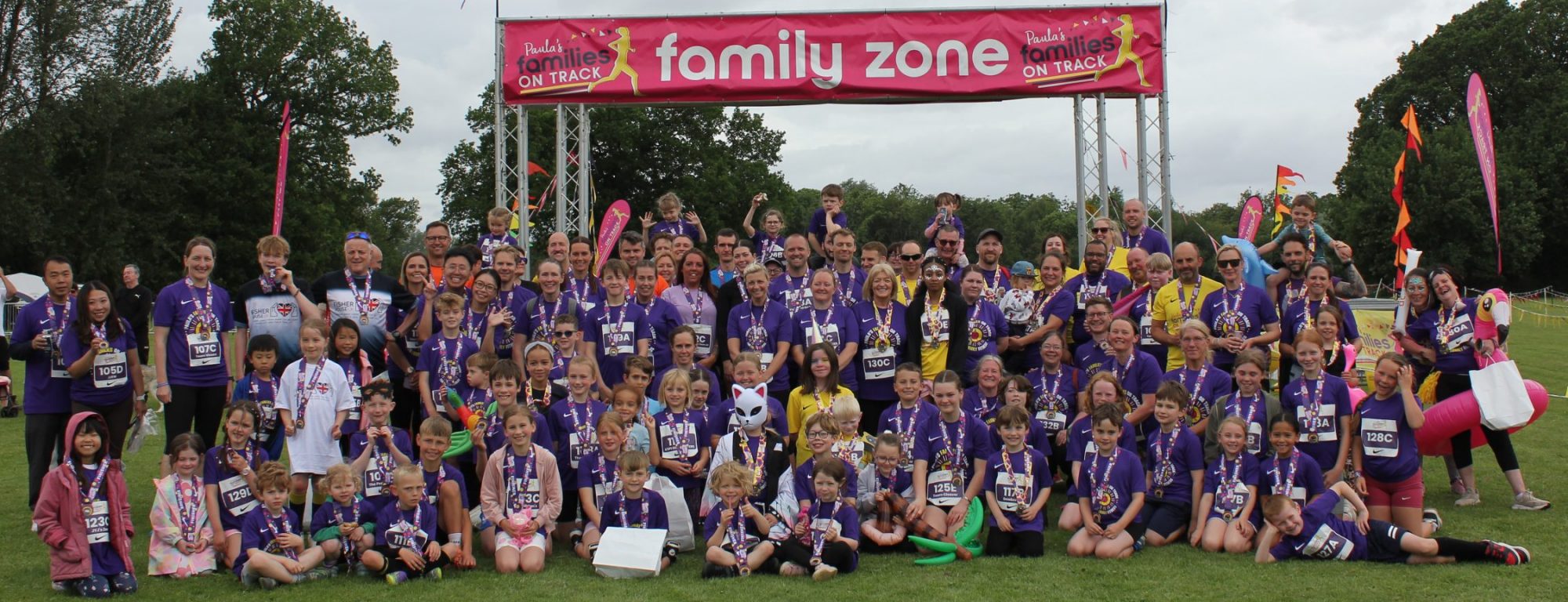 Large group of adults and children wearing matching purple T‑shirts and medals pose on grass beneath a pink “family zone” banner at an outdoor event, with rainbow flags, trees, and an overcast sky in the background.